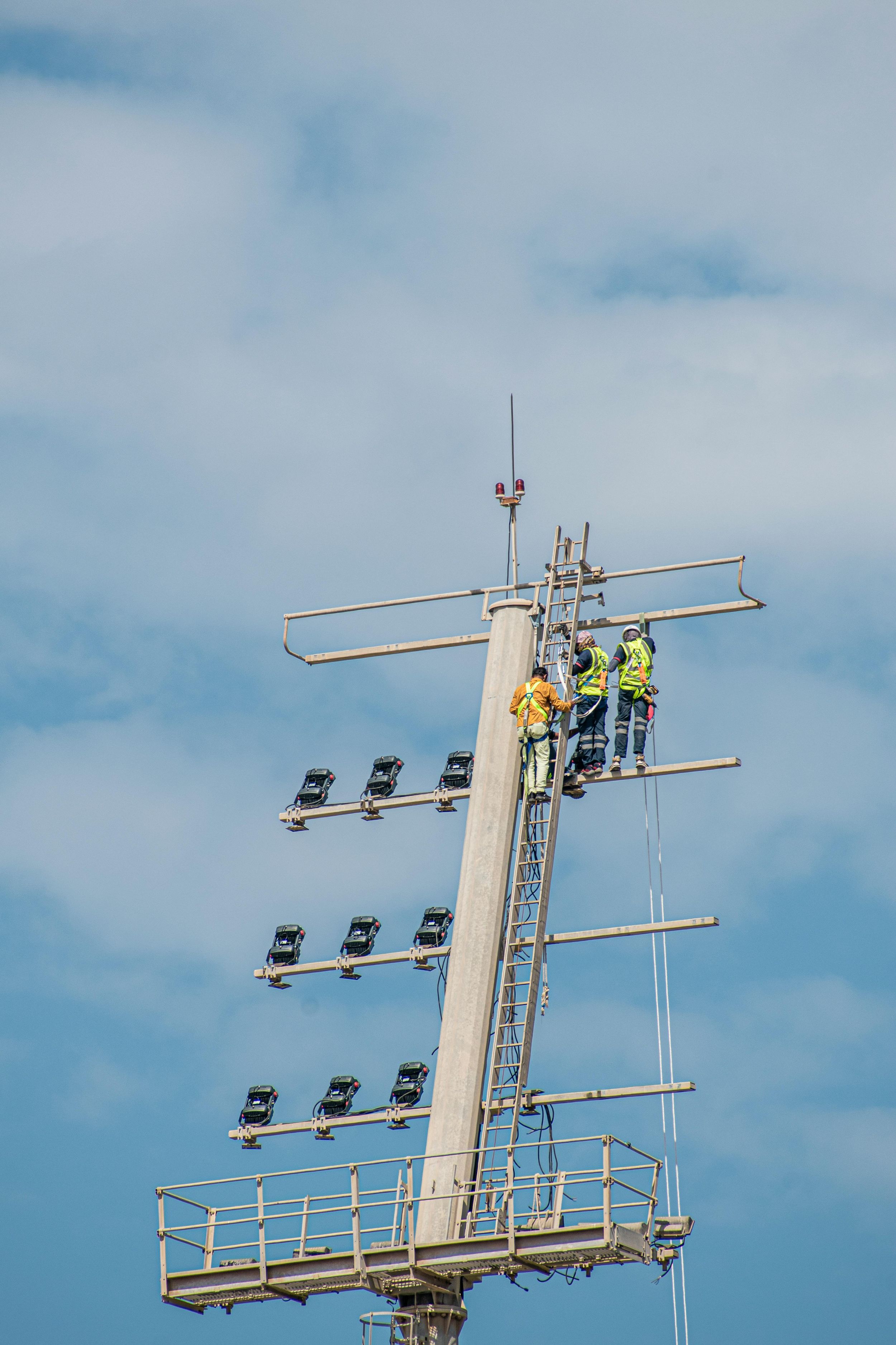 Workers performing maintenance on a tall signal pole with multiple light fixtures against blue sky.