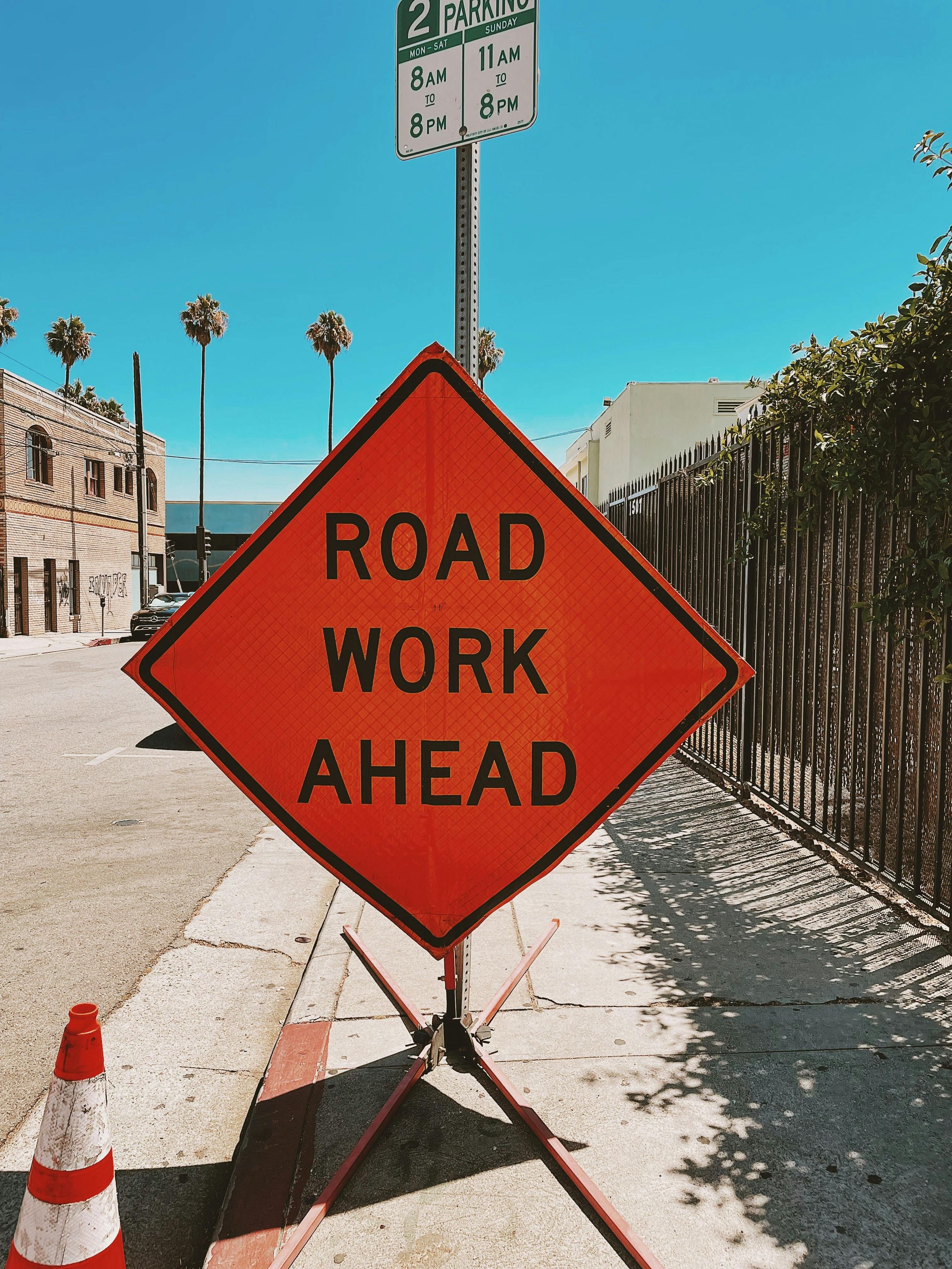 Orange diamond-shaped road work ahead sign on sidewalk with palm trees and buildings in background.