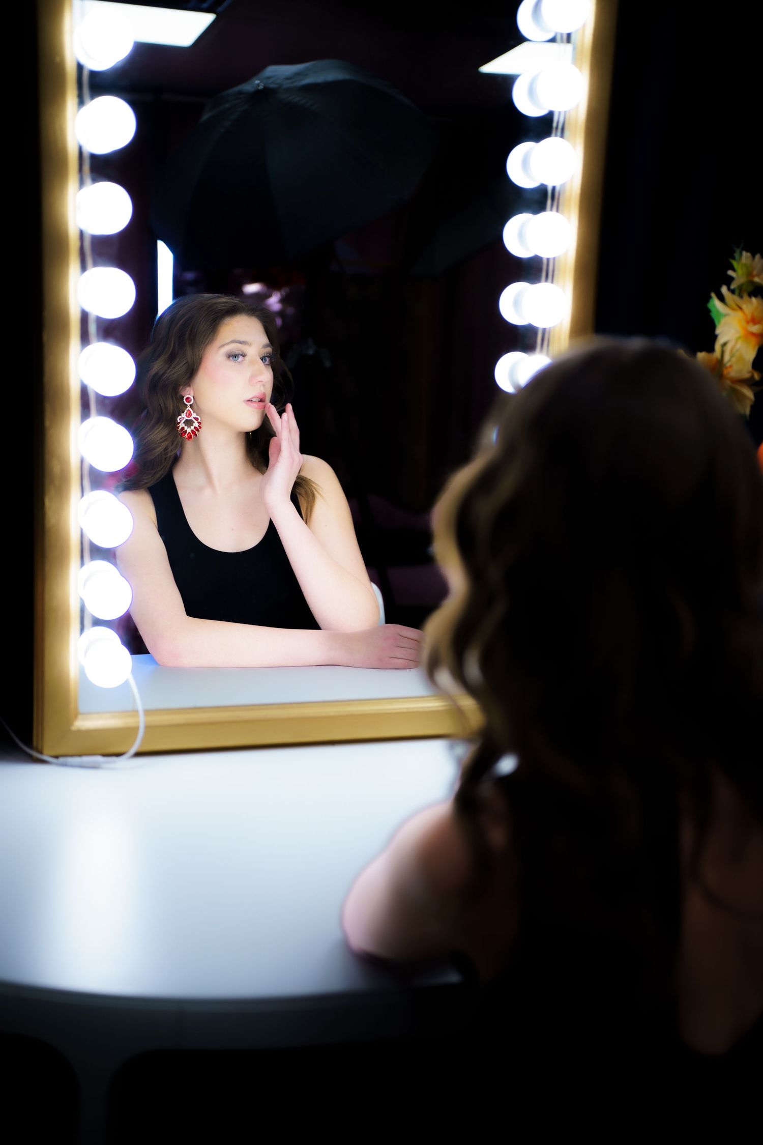 Reflection in illuminated makeup mirror shows silhouette wearing black tank top with dramatic lighting and vanity lights.