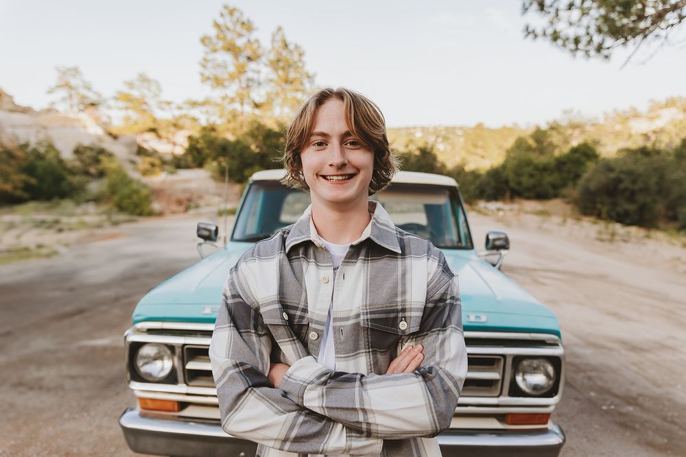 Smiling portrait in front of vintage turquoise truck wearing plaid jacket with arms crossed.