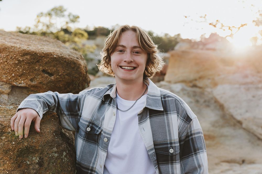 Natural sunlit portrait near rock formations wearing grey plaid jacket and white t-shirt.