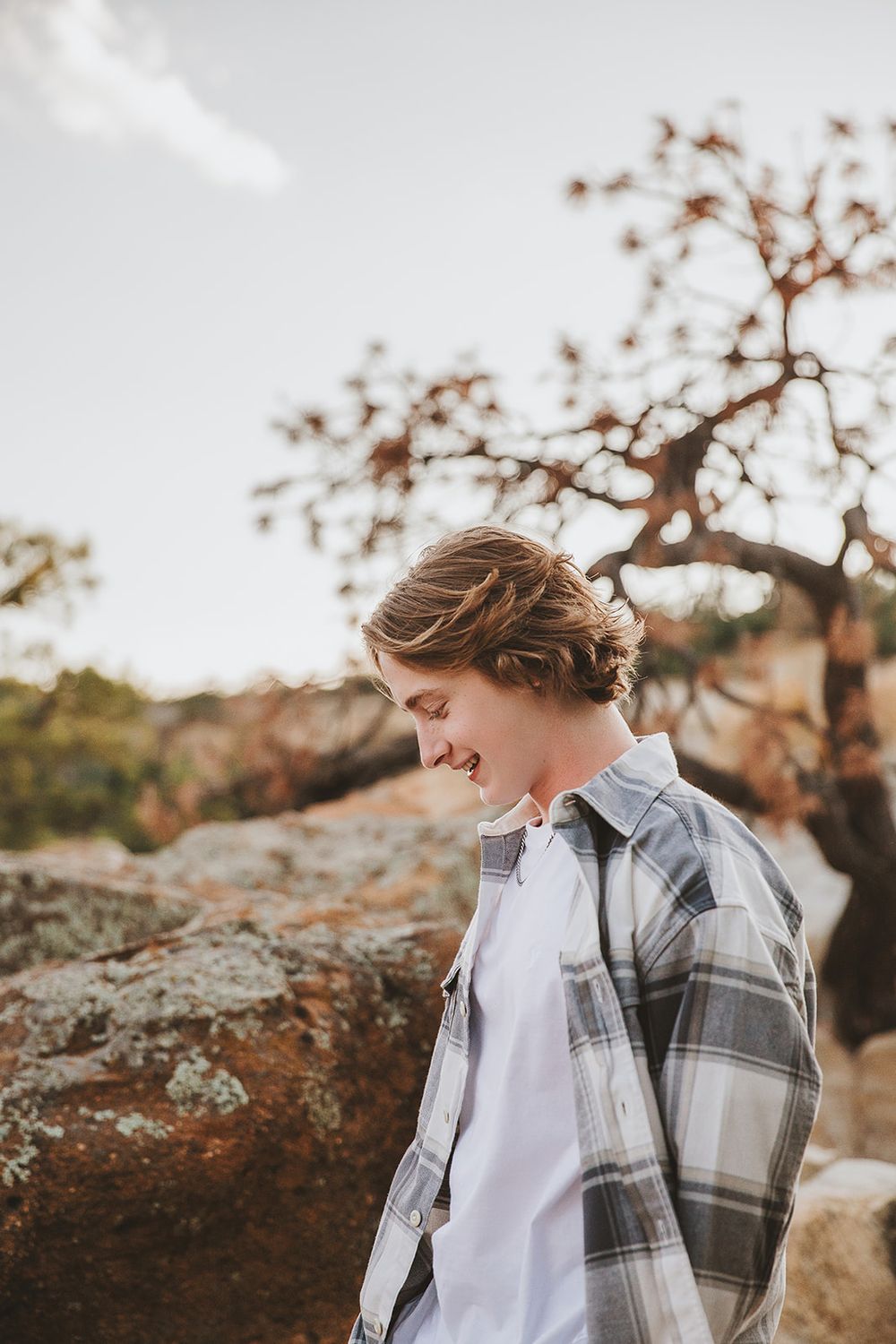 Candid portrait at sunset showing joyful expression while wearing grey plaid shirt against rocky backdrop.