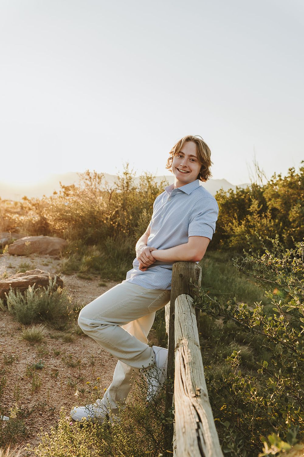 Relaxed pose leaning on wooden fence during sunset wearing light blue polo and jeans.