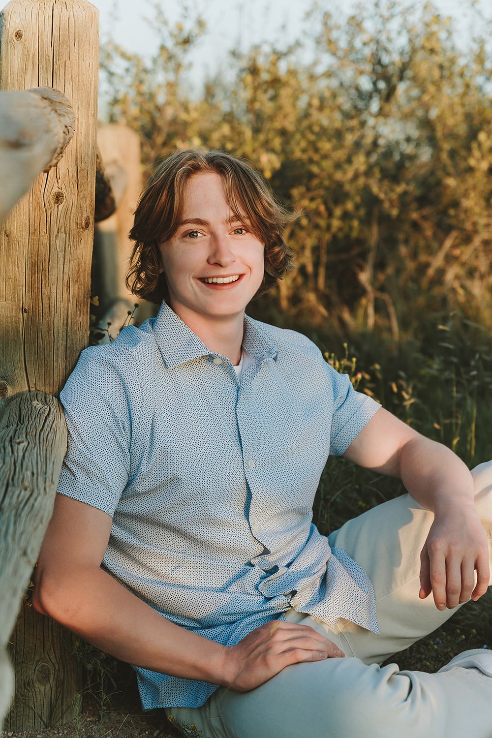 Natural outdoor portrait beside wooden post wearing light blue patterned shirt at golden hour.
