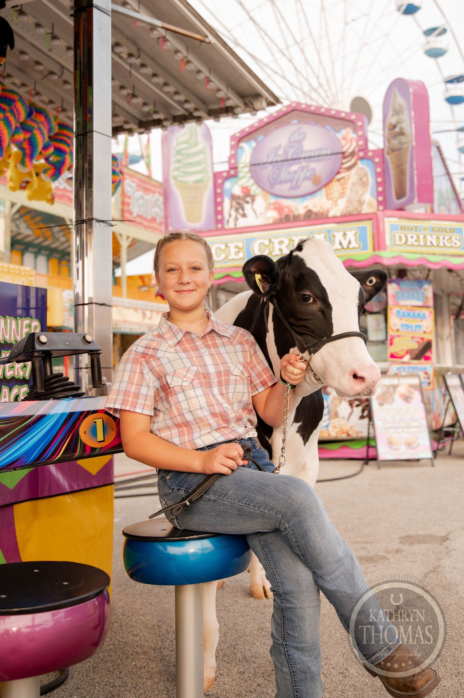 Young exhibitor sitting with dairy calf at colorful carnival fairgrounds.