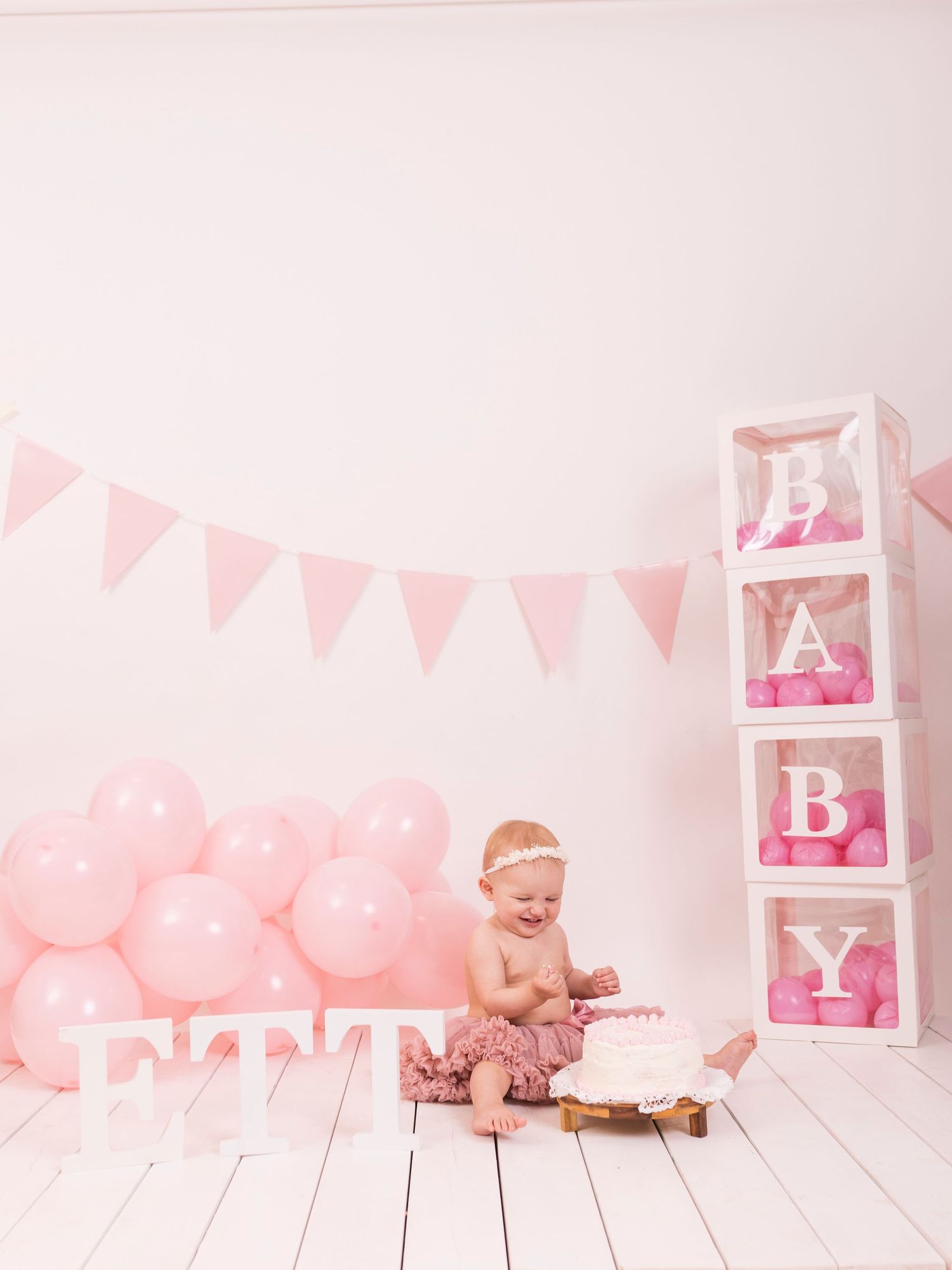 Baby wearing pink tutu skirt laughs while standing next to cake and pink balloons during first birthday photoshoot.