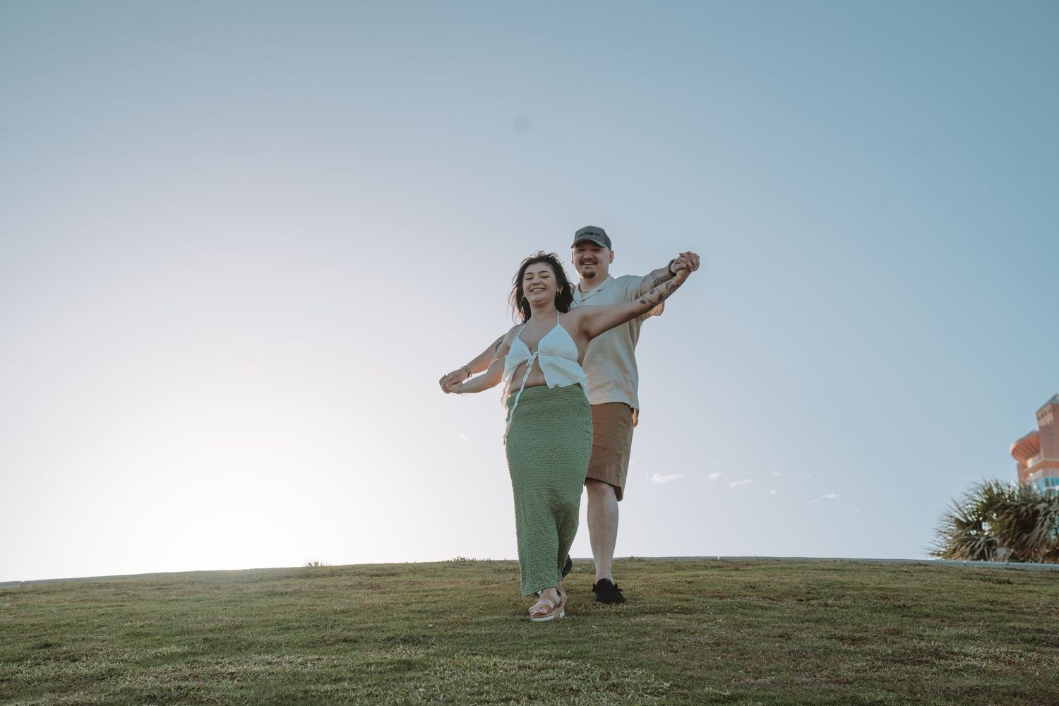Two people stand together on a grassy hilltop against a bright sky, posing with their arms outstretched.