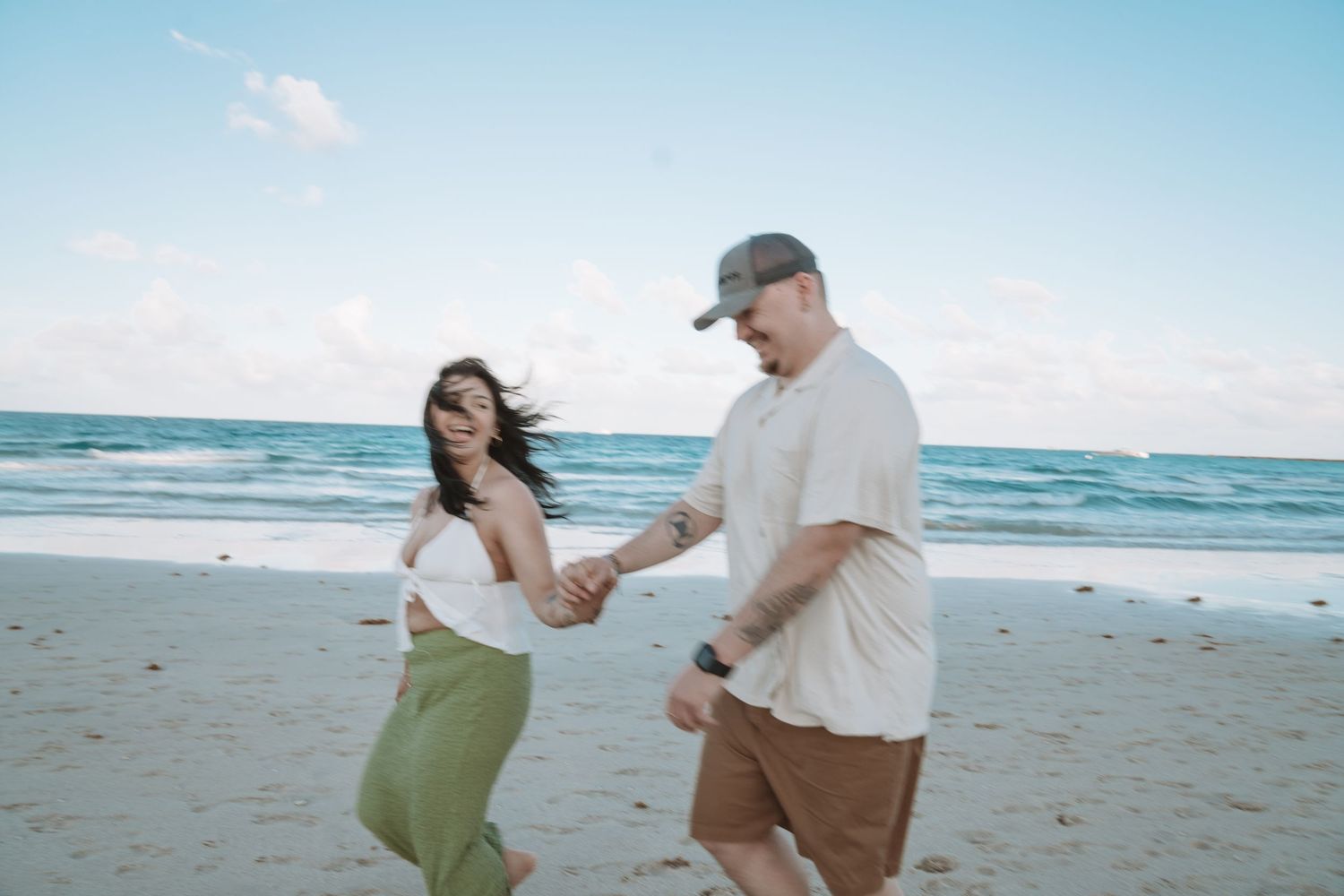 A couple holding hands and smiling while walking together on a sunny beach with ocean waves in the background.