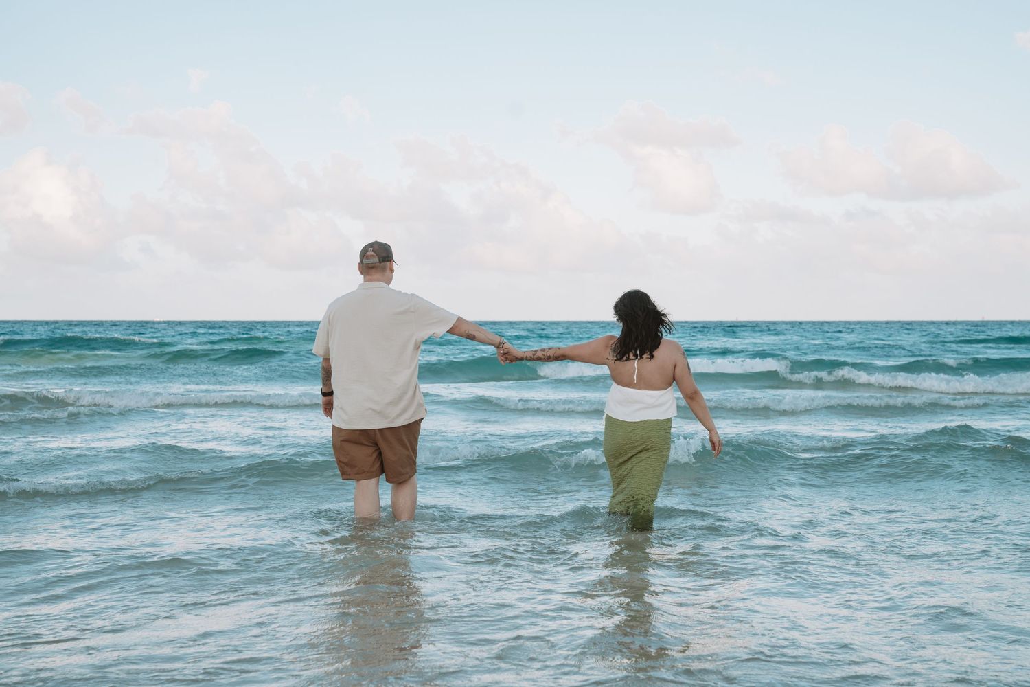 Two people wade together in turquoise ocean waters at the beach under a soft cloudy sky.
