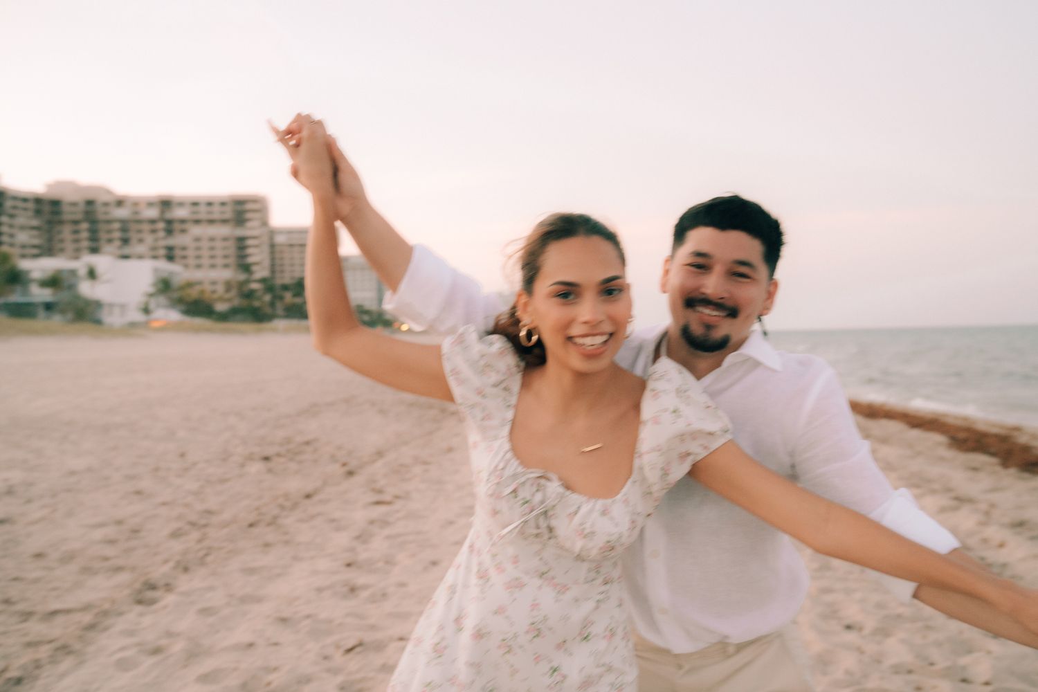 A happy couple wearing white outfits dances and poses together on a sandy beach at sunset.