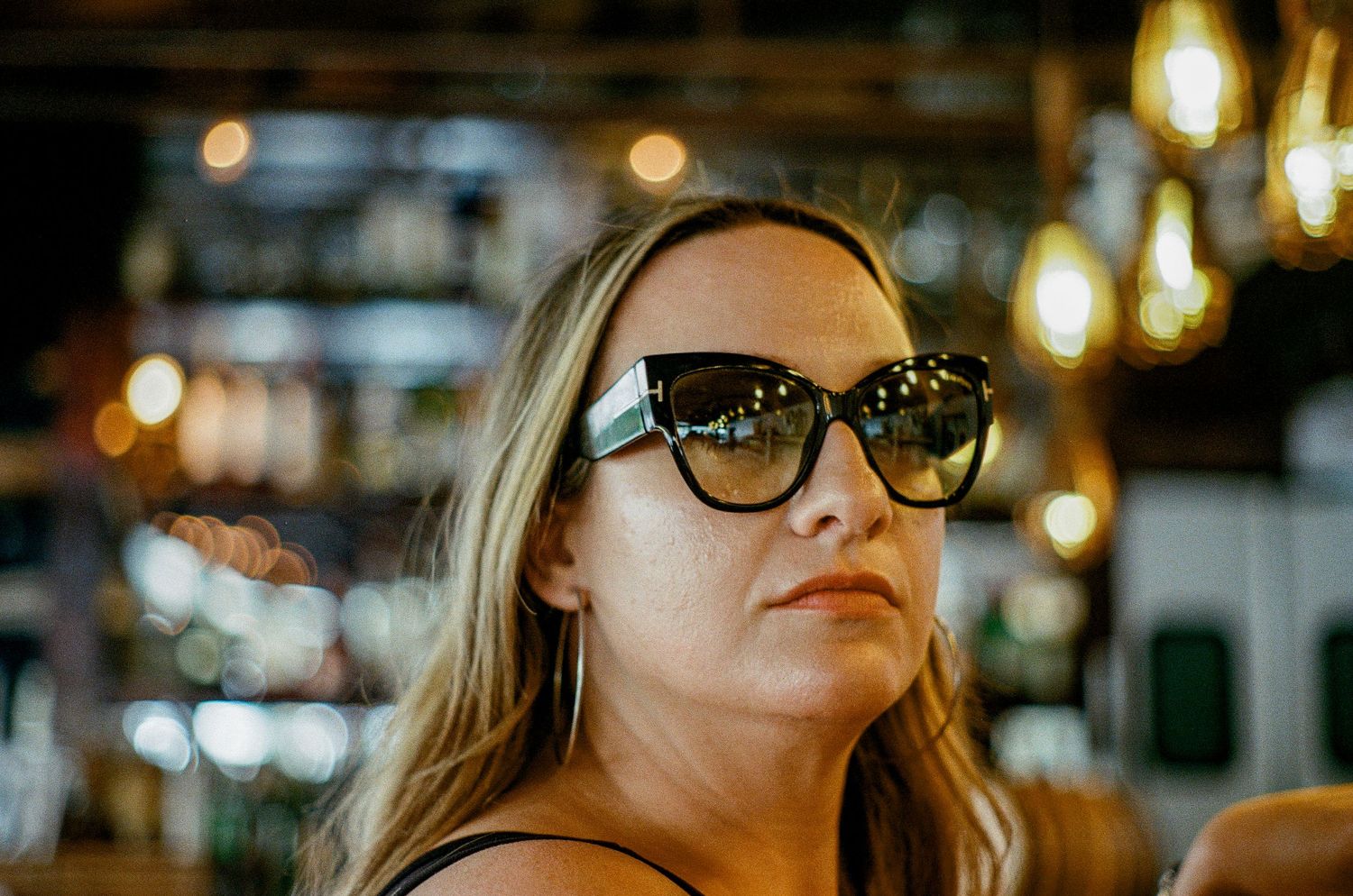 A woman with long blonde hair wearing sunglasses looks up in a bar with bokeh lighting in the background.