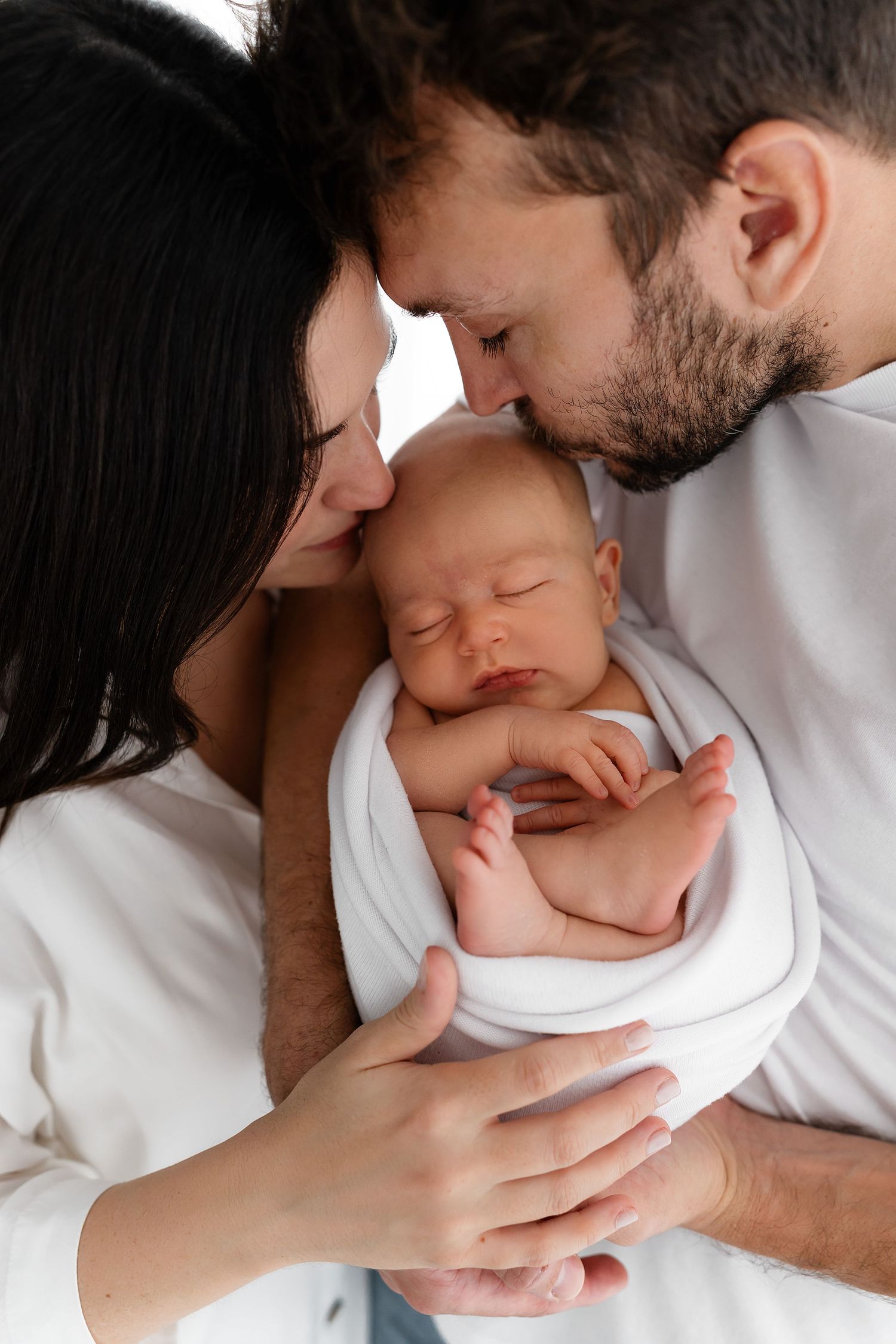 A baby sleeps peacefully swaddled in white while being lovingly held in their parent's arms.