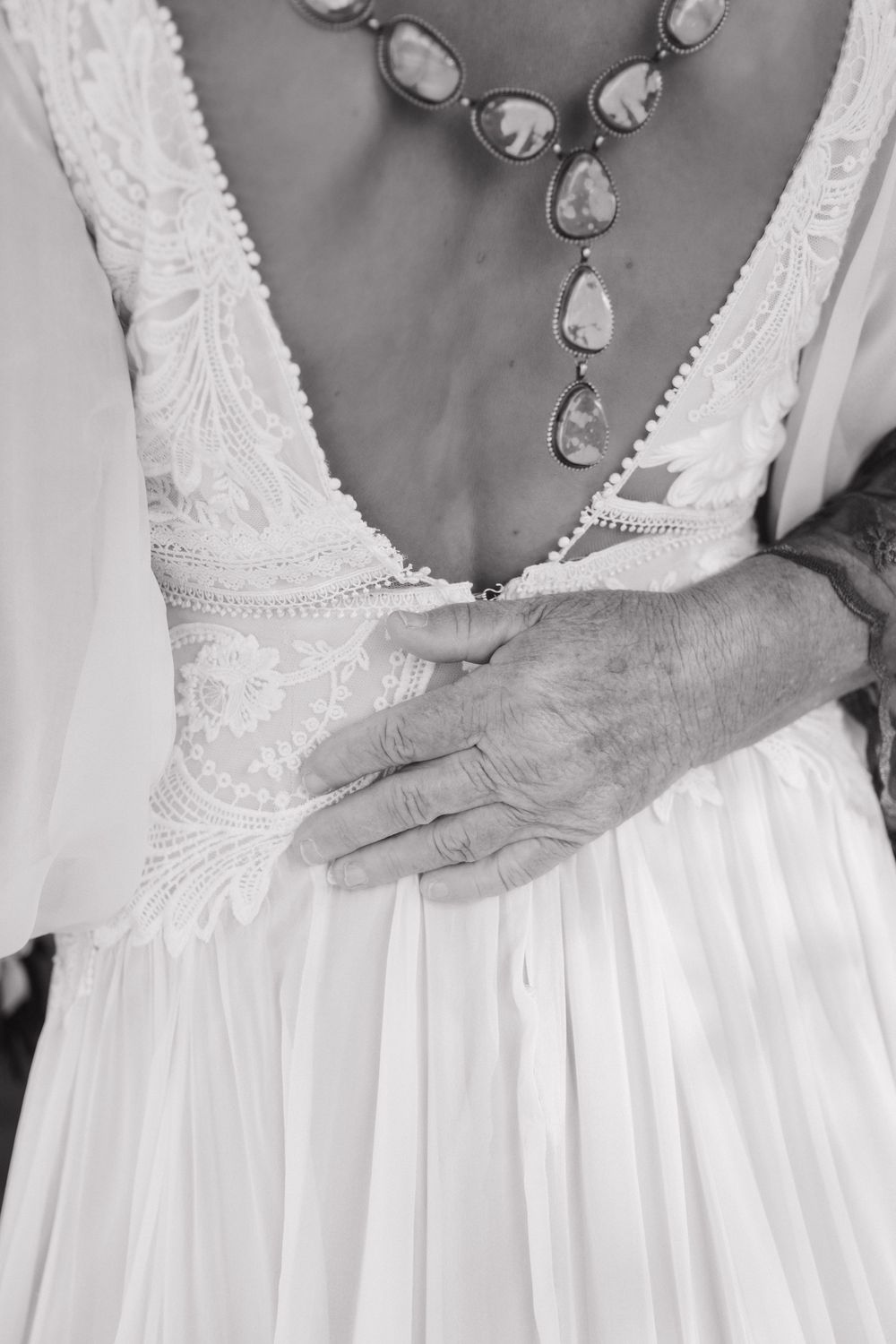 A close-up black and white photo of a white lace wedding dress bodice with a statement necklace and hands adjusting the back.