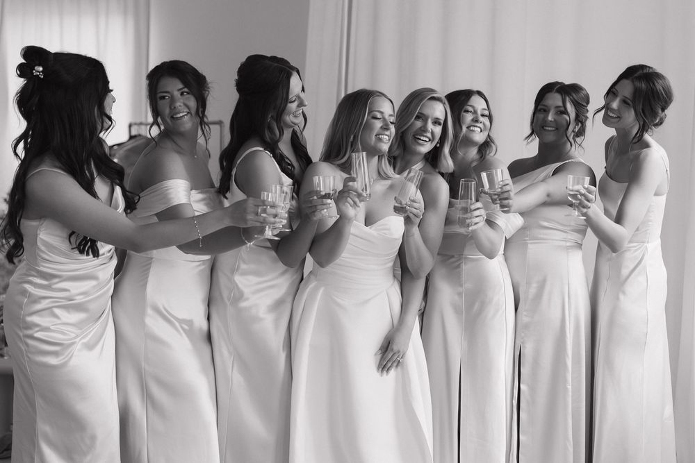 Black and white photo of bridesmaids in white gowns laughing and toasting with champagne glasses at a wedding.