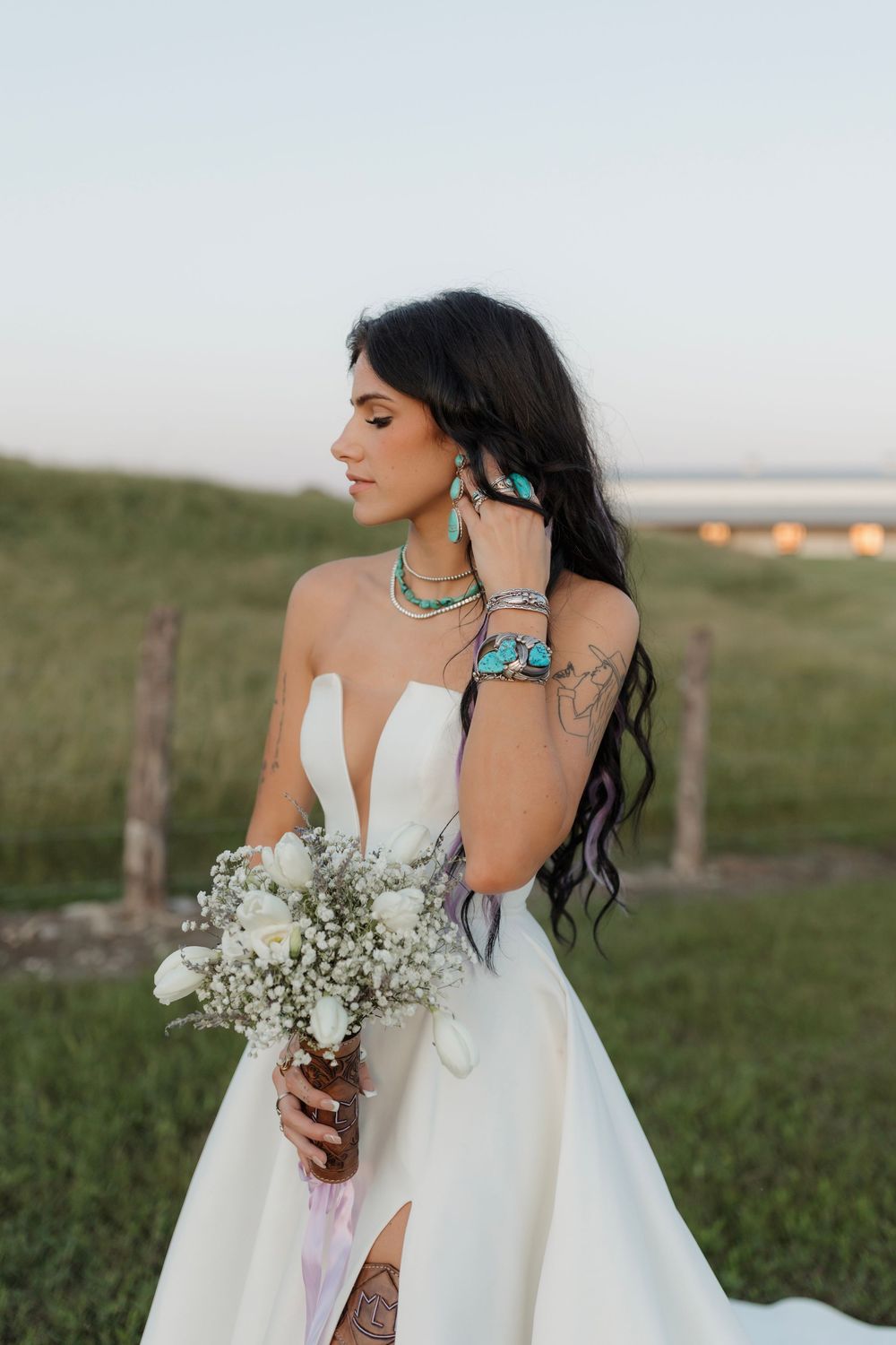 Bride in white strapless gown with turquoise jewelry holds white bouquet outdoors in green field at dusk.