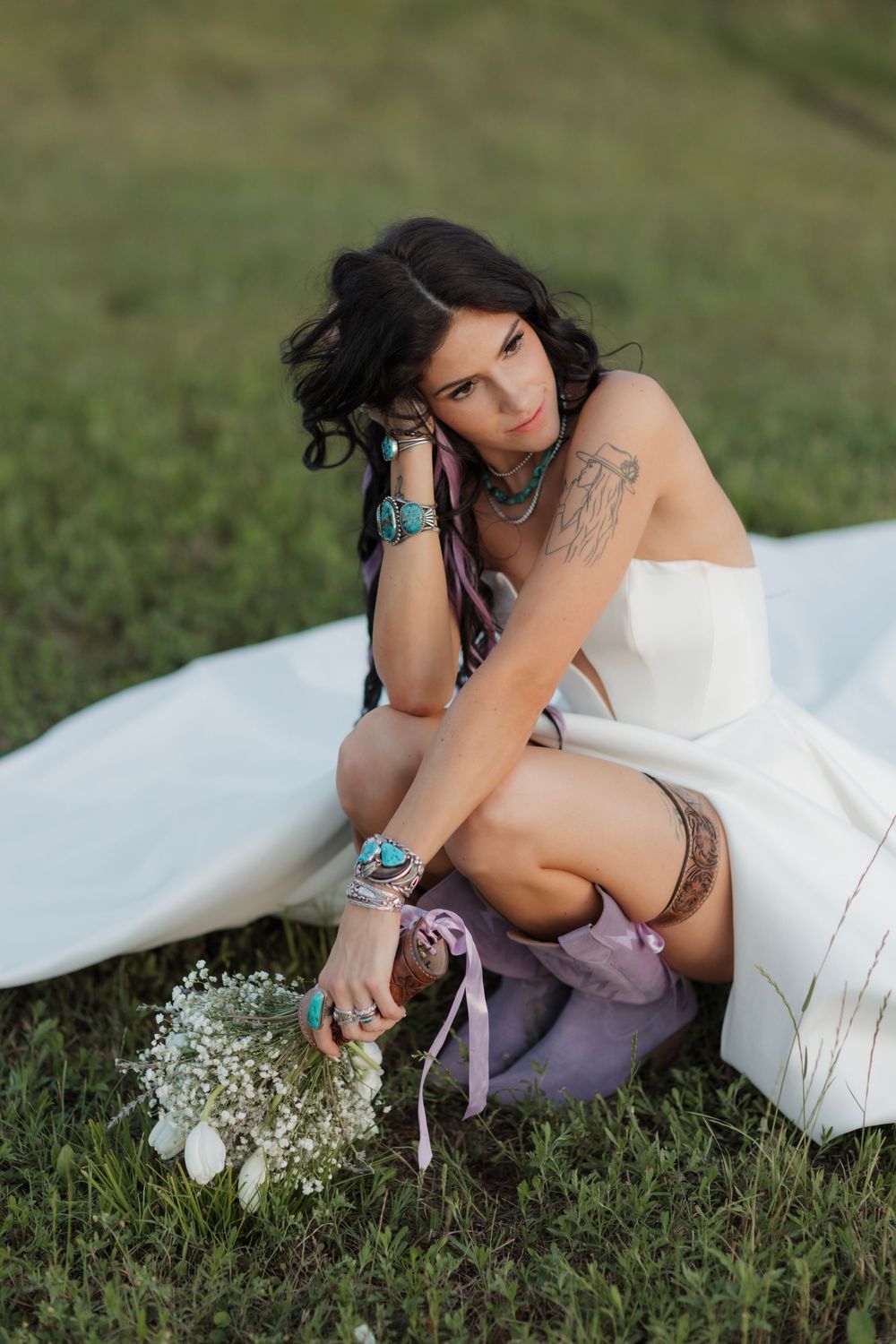 Bohemian bride in white strapless gown sitting on grass with turquoise jewelry and small white floral bouquet.