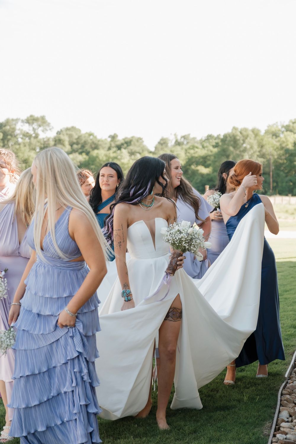 Bride in a white gown surrounded by bridesmaids in blue dresses celebrating outdoors at a wedding.