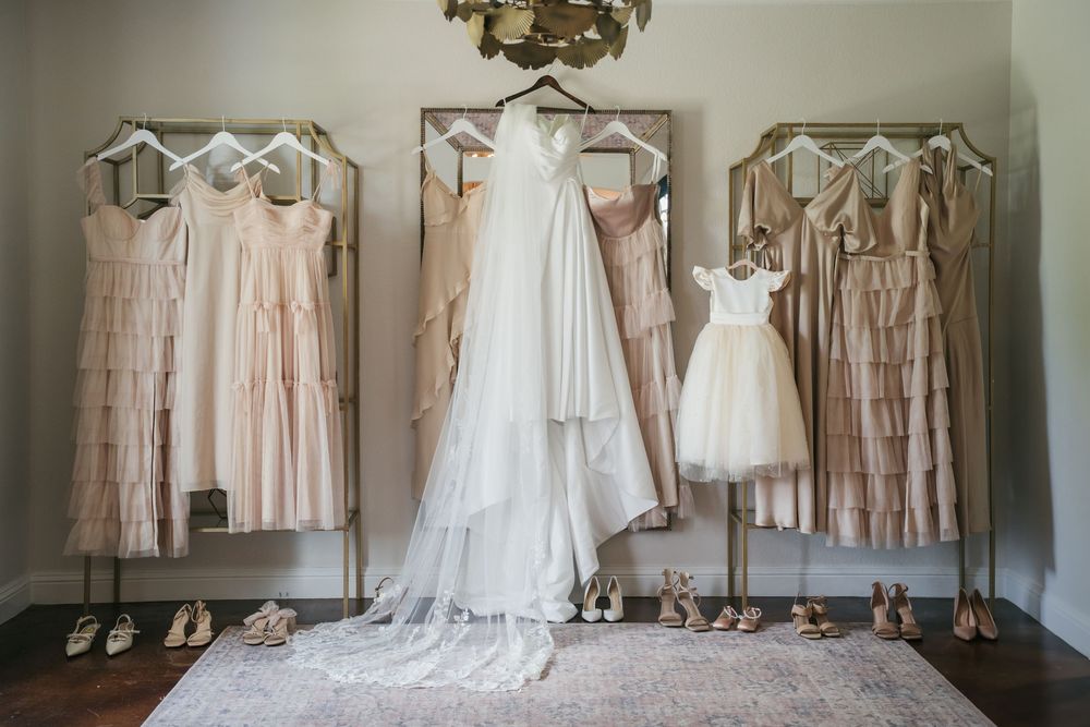 Wedding gowns and bridesmaids dresses in blush pink and white hang on elegant display racks with matching shoes below.