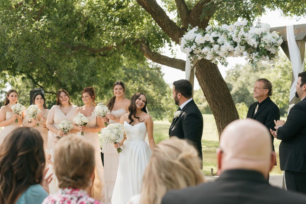 A wedding ceremony takes place outdoors beneath a large shady tree with guests looking on.
