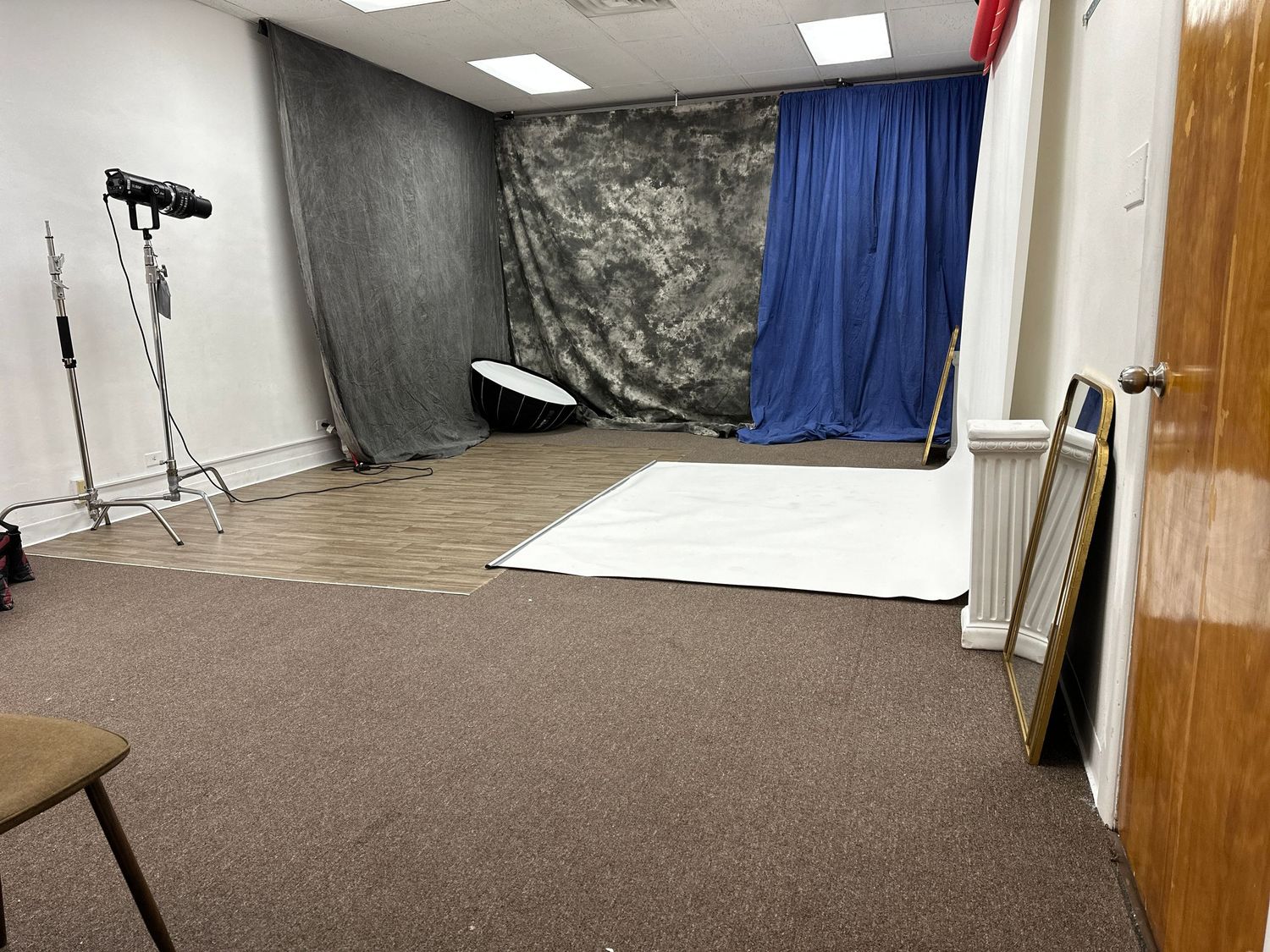 Small photo studio setup with brown carpet, backdrop stands, and gray and blue backdrop curtains in corner of room.