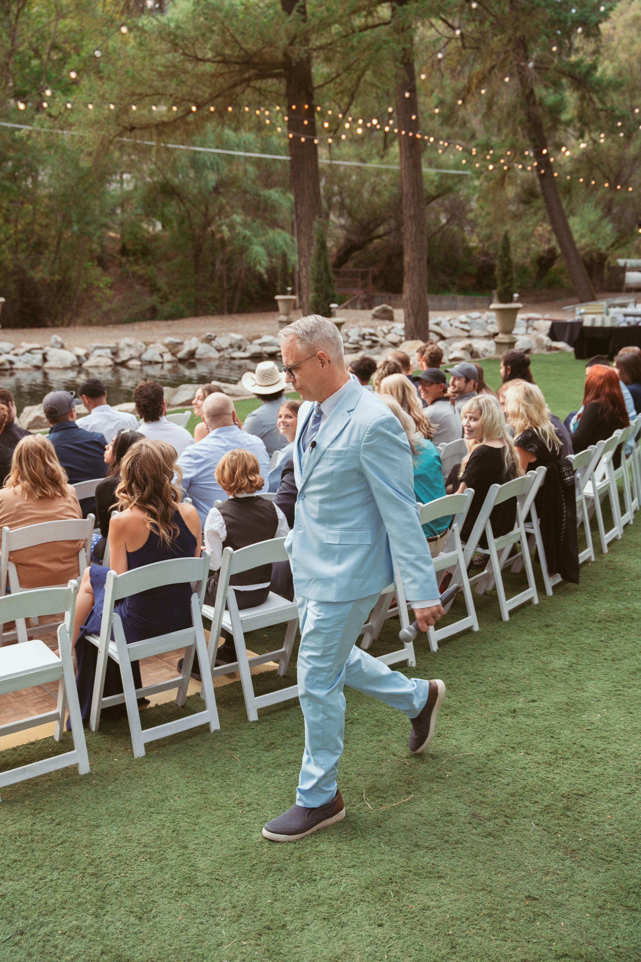Outdoor wedding guest in light blue suit walks past rows of white folding chairs under string lights.