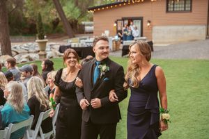 Wedding guests walking down an outdoor ceremony aisle at a rustic venue with string lights and grass lawn.