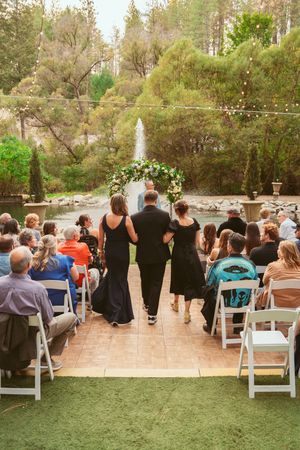 An outdoor wedding ceremony with guests seated in white chairs and a floral arch backdrop in a wooded setting.