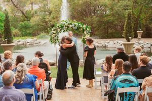 An outdoor wedding ceremony takes place under a floral arch with chairs arranged for guests on a stone patio area.
