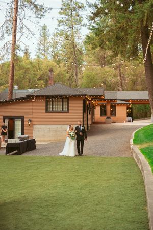A wedding couple stands outside a rustic brown venue surrounded by pine trees and string lights.