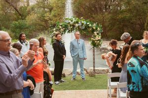 Wedding guests gather around a floral arch during an outdoor ceremony with water feature in background.