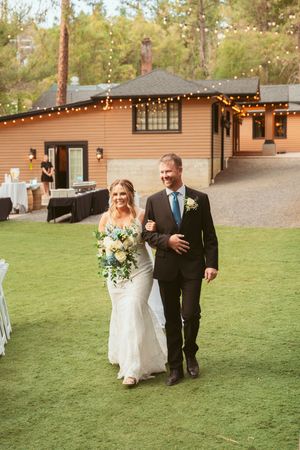 Couple walks together on a grassy lawn at an outdoor wedding ceremony with string lights hanging above.