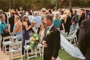 Wedding guests mingling after outdoor ceremony with white chairs and golden sunlight filtering through trees.