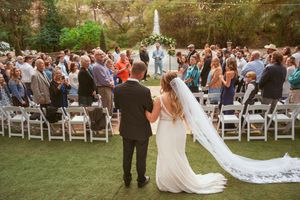 Outdoor garden wedding ceremony with white chairs and guests gathered around a floral arch during sunset.