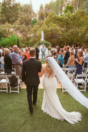 A wedding ceremony takes place outdoors with a long white veil trailing behind as guests watch from white chairs.