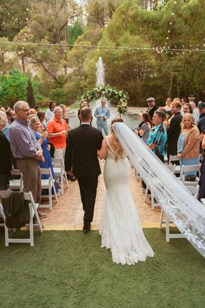 Wedding ceremony scene with string lights overhead and white chairs set up on a patio surrounded by trees.