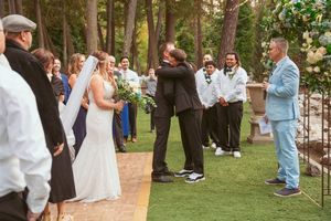 An outdoor wedding ceremony takes place on artificial turf surrounded by tall pine trees with guests gathered around.