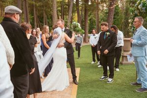 An outdoor wedding ceremony takes place on a grassy lawn surrounded by tall evergreen trees during a sunny day.