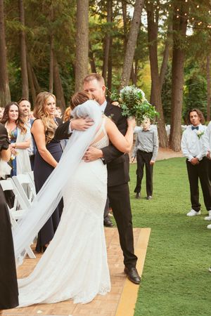 Wedding celebration scene with guests on a grassy lawn surrounded by tall trees.