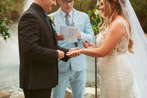 A wedding ceremony under a floral arch as couple exchanges rings with officiant in light blue suit.