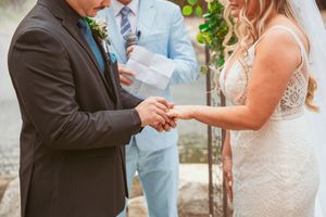 Couple exchanging wedding rings during outdoor ceremony with officiant in light blue suit.