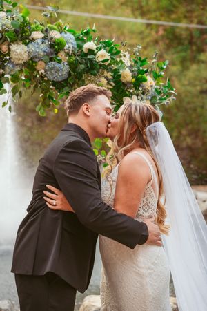 Romantic wedding couple shares their first kiss under a beautiful floral arch with hydrangeas and roses.