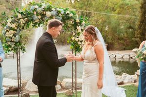 An exchange of rings takes place under a beautiful floral arch at an outdoor wedding ceremony.