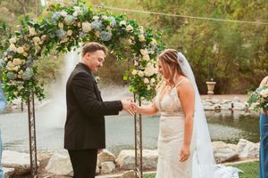 A joyful moment during ring exchange under an elegant white floral arch at an outdoor ceremony.