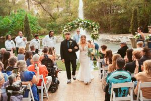 A wedding recessional down an outdoor aisle with guests seated on white chairs and a waterfall in the background.