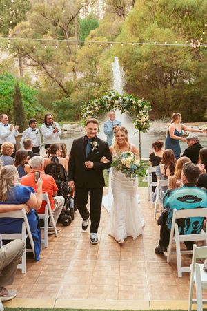 Wedding ceremony processional on a stone patio with guests seated in white chairs under string lights.