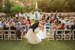 A celebratory dip pose during a wedding ceremony with guests seated on white chairs outdoors.