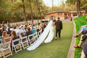 Outdoor wedding venue with rows of white chairs set up on grass under tall trees.