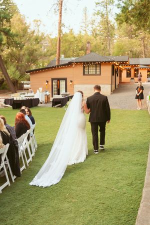 A long white wedding veil trails behind as couple walks on grass path near rustic venue building.