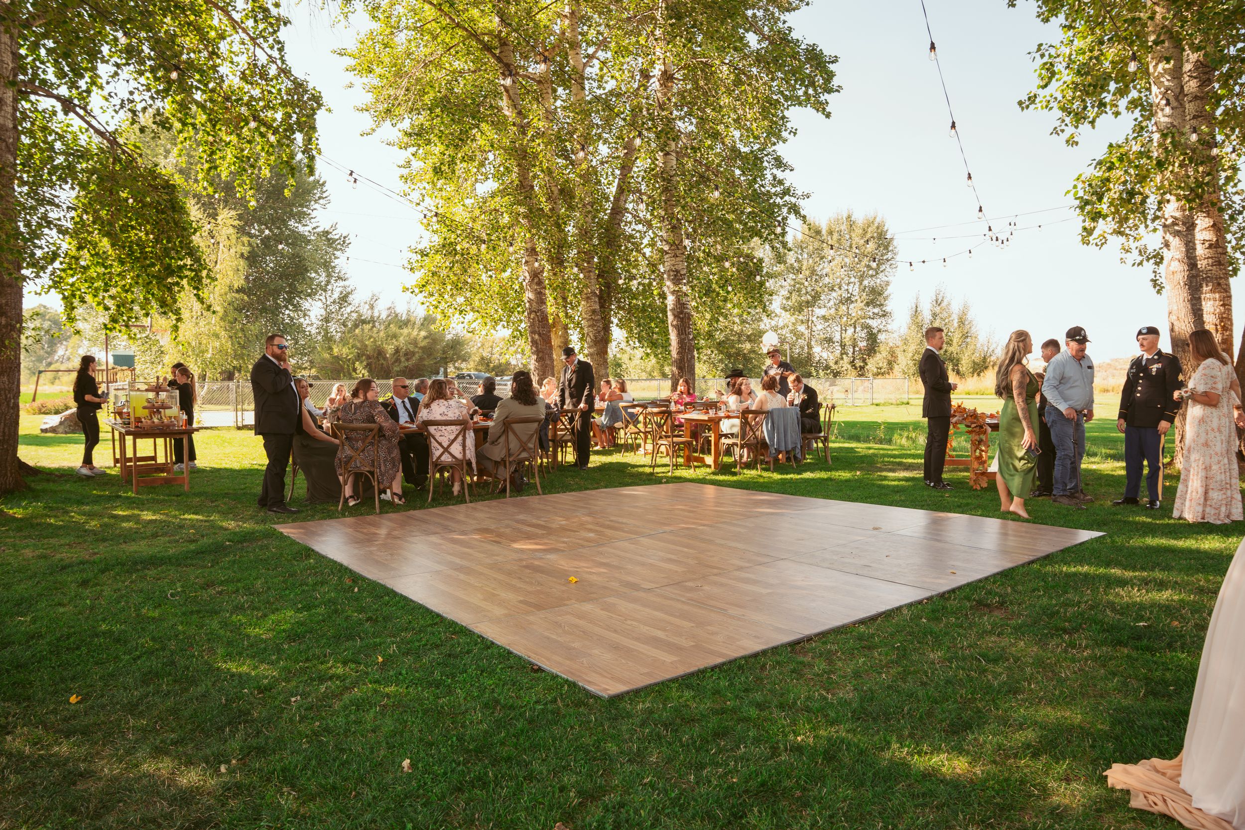 A wooden dance floor is set up outdoors on grass for a garden party with guests gathered around under tall trees.