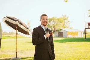 Man in formal black suit stands on sunny lawn speaking into microphone at outdoor event.
